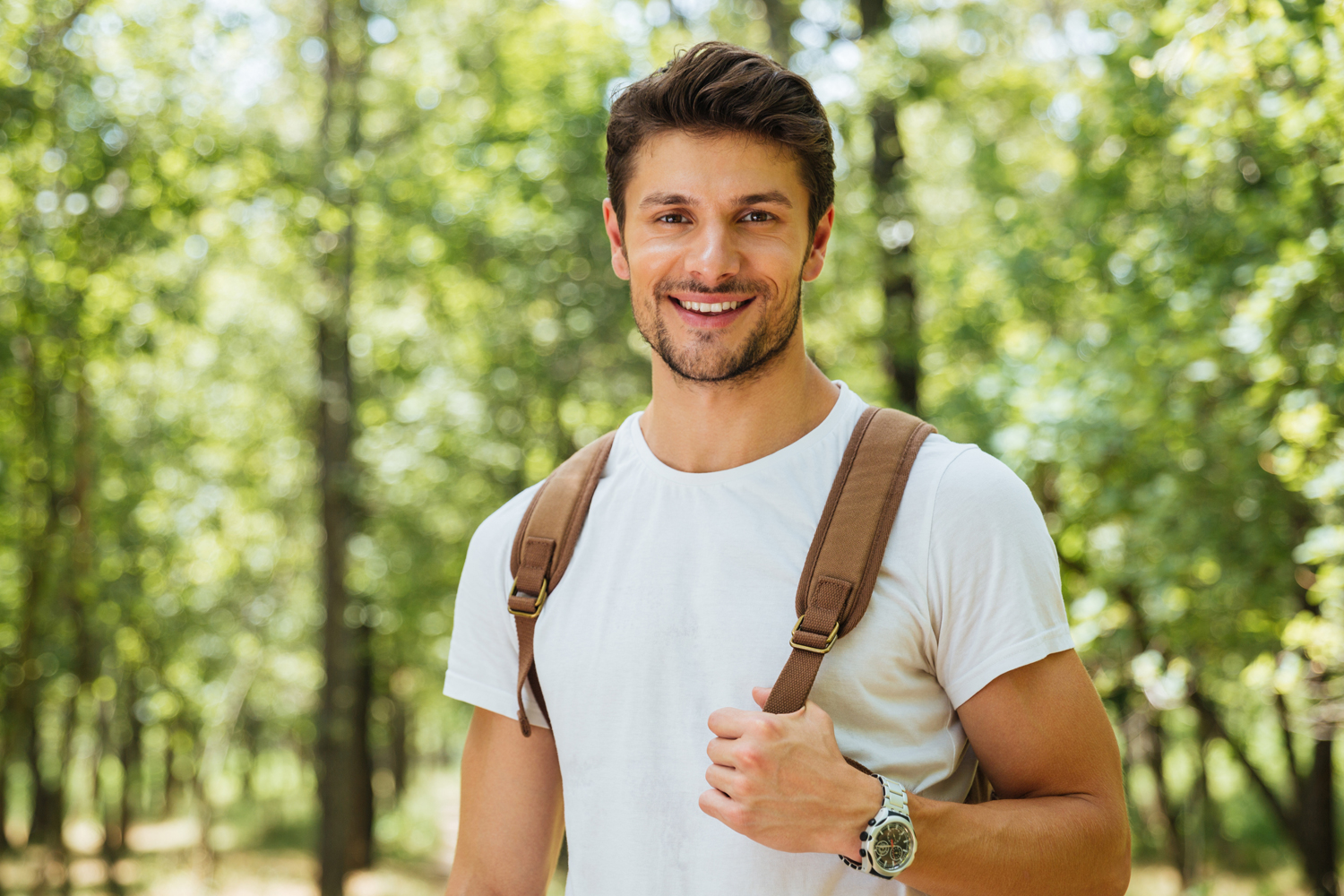 A young man standing outside with a brown backpack on, smiling at the camera